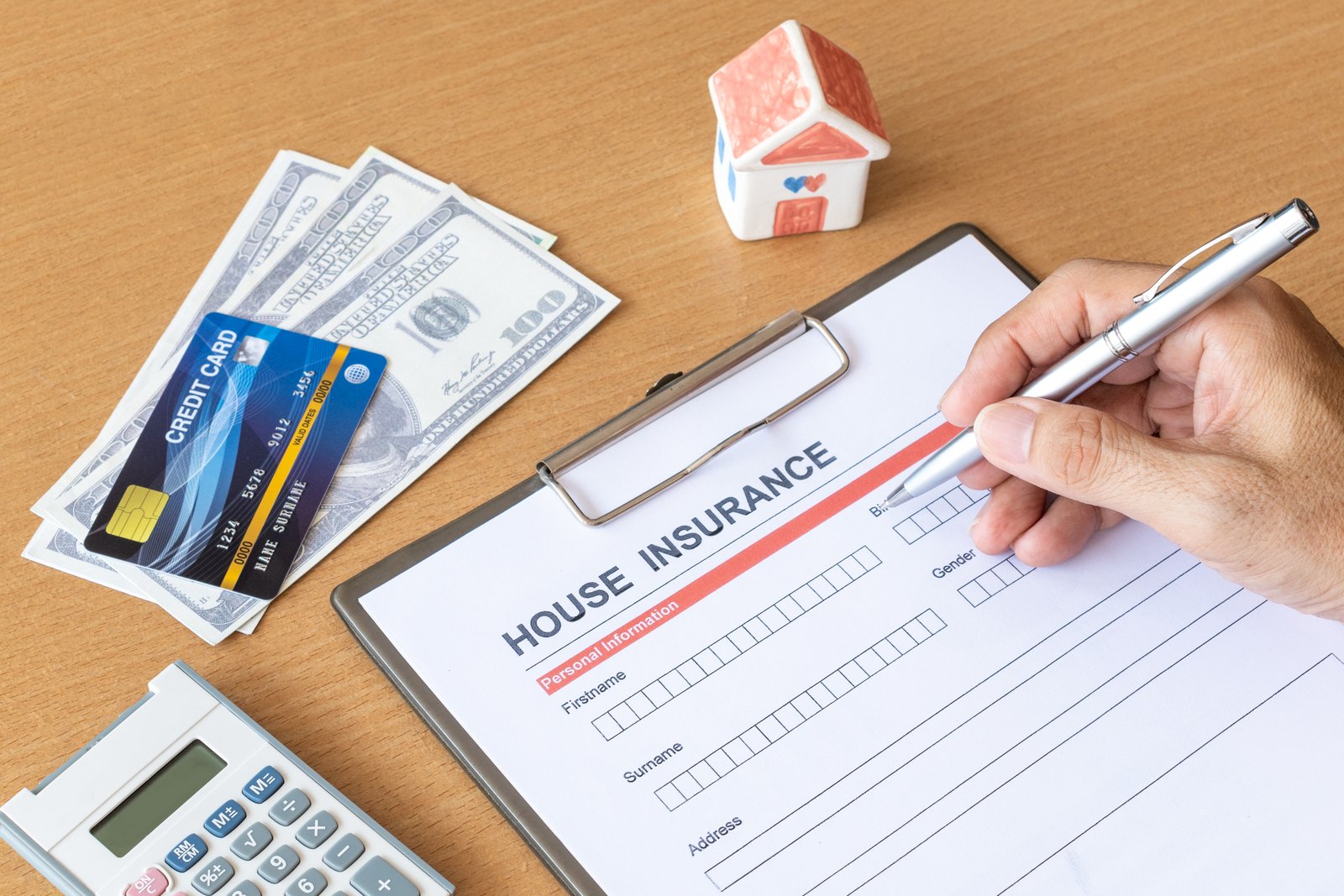 A hand filling out a printed House Insurance application form on a clipboard, next to cash, a credit card, a calculator, and a small toy house